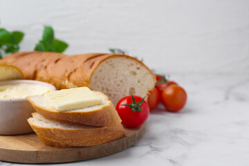 Cut baguette with butter and tomatoes on white marble table, closeup. Space for text