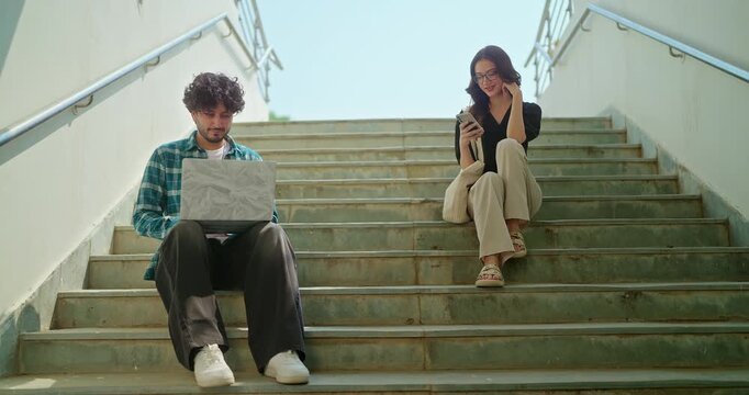 Indian young adult gen z busy male man use laptop look screen do study check online e work at campus place. Happy female sitting on stair hold mobile phone read notes enjoy new day spend break time