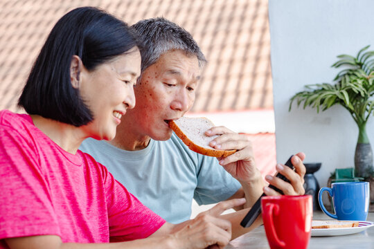 A middle-aged Asian couple enjoys their morning coffee and breakfast on a balcony together. The woman scrolls on her mobile phone while her husband eats toast and looks at her phone