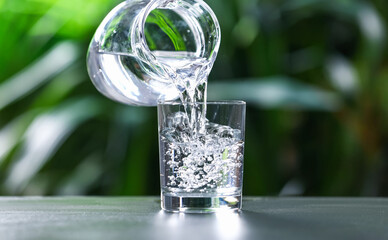 Pouring water into glass from jug at grey table against blurred green background, closeup