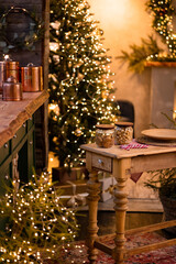 A cozy room with Christmas trees decorated with garlands. A table with dishes and decorative elements can be seen.