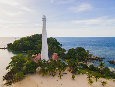 Stunning aerial view of Lengkuas Island, Belitung, Indonesia, at sunset. The iconic white lighthouse stands amidst granite boulders and wooden boats, surrounded by clear turquoise water.
