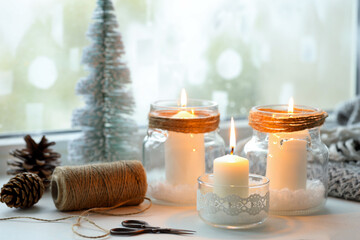 DIY Christmas lanterns. Burning candles in glass jars, cones, twine and scissors on window sill indoors, closeup