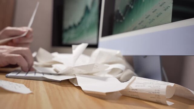 The video pans across a cluttered desk with receipts in front of dual monitors showing a rising chart. Ideal for concepts of accounting, finance growth and audits.