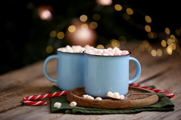 Tasty cocoa with marshmallows and candy canes on wooden table against blurred Christmas lights, closeup