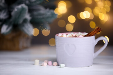 Tasty cocoa with marshmallows and cinnamon on white wooden table against blurred Christmas lights, closeup