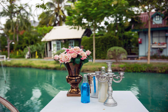 A beautiful romantic outdoor setting featuring a vase of pink roses, a silver candelabra, and vintage bottles arranged on a white table, quaint rustic cottages, ideal for wedding receptions - Powered by Adobe