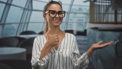 Young woman wearing glasses and striped blouse points finger to open palm in building restaurant interior while smiling and offering gesture; confidence.