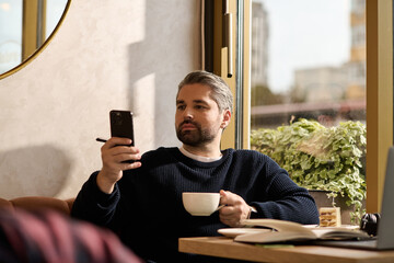 Handsome mature man enjoys coffee while engaging with his phone in a cozy cafe setting