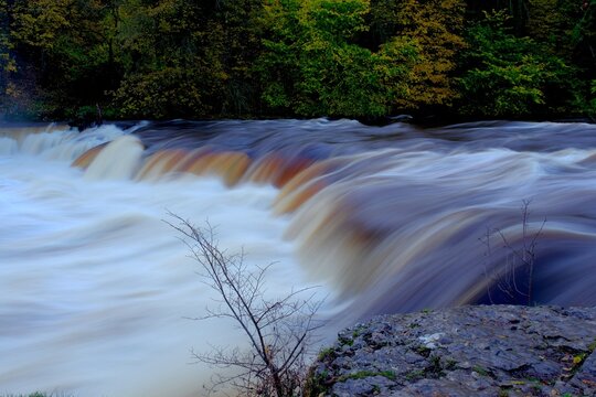 Long-exposure autumn waterfall with smooth, rushing water cascading over amber-tinted ledges. Bare branches and wet rock frame the river, with colourful woodland in the background. - Powered by Adobe