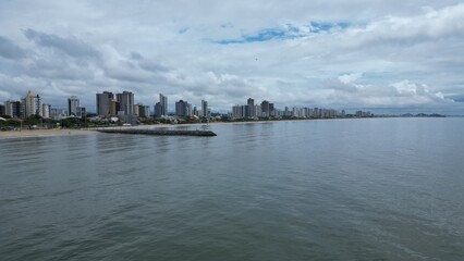 Aerial shot of a long pier jutting into a calm, gray sea under a cloudy sky. Unidentifiable people stand at the end, with distant islands on the horizon. Peaceful, minimalist coastal scene.