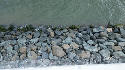 Top view of a rocky seawall with mossy stones, gentle waves, and a white concrete walkway along the coast.