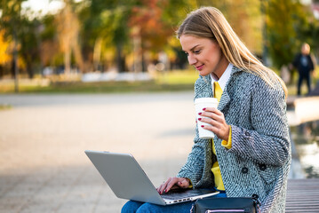 Portrait of modern businesswoman in autumn coat using laptop and drinking coffee while sitting on a park bench in the city. 