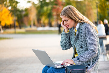 Portrait of overworked and stressed businesswoman with headache using laptop while sitting on a park bench in the city.