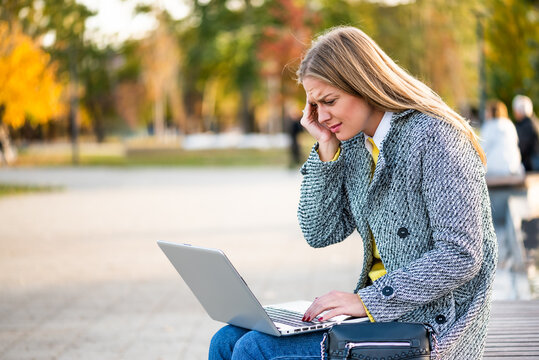 Portrait of overworked and stressed businesswoman with headache using laptop while sitting on a park bench in the city. - Powered by Adobe