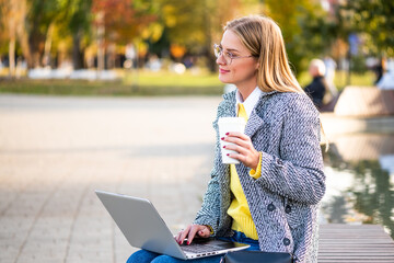 Portrait of urban businesswoman with eyeglasses in autumn coat using laptop and drinking coffee while sitting on a park bench in the city. 