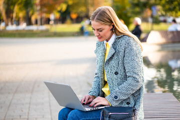 Portrait of urban businesswoman in autumn coat using earphones and laptop while sitting on a park bench in the city. She is having a video call.	