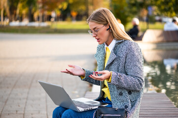 Portrait of urban businesswoman with eyeglasses in coat using earphones and laptop while sitting on a bench in the city park. She is talking and gesturing during a video call.	