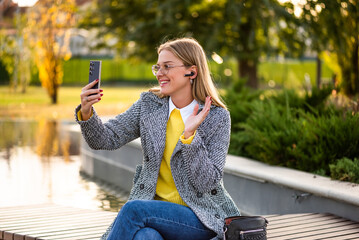 Portrait of urban businesswoman with eyeglasses in stylish autumn coat wearing earphones and waving during a video call on her smartphone while sitting on park bench in the city.	