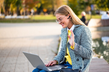 Portrait of stylish businesswoman with eyeglasses in coat wearing earphones and waving during a video call on her laptop while sitting on park bench in the city during sunny autumn day.