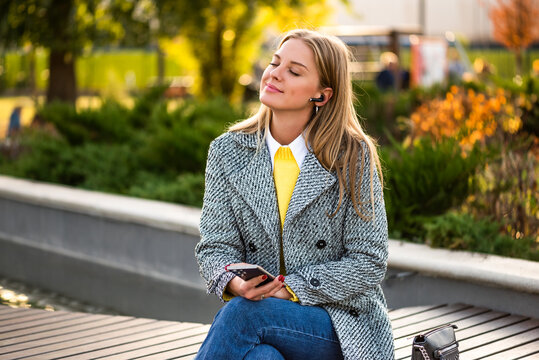 Portrait of beautiful urban businesswoman in autumn coat listening to music through earphones and using smartphone while relaxing on a park bench in the city.