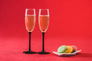 Two champagne flutes filled with bubbly liquid sit beside a small dish of colorful macarons on a vibrant red background