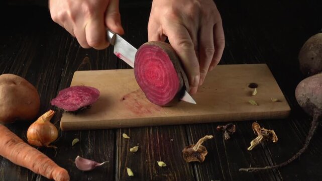 A person skillfully slices a vibrant beet on a wooden cutting board. Nearby are fresh carrots, onions, and various herbs, creating a lively kitchen scene filled with fresh produce.