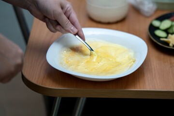 Mixing eggs and milk with a metal fork in a white ceramic bowl to make an omelet