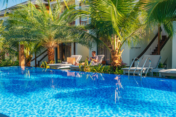 Man with smartphone relaxing on sun lounger by the swimming pool at luxury hotel surrounded by palm trees on Phu Quoc island, Vietnam. Tourist resting in topical resort on summer vacation in Asia