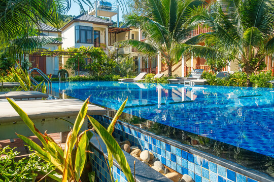 Blue swimming pool with sun loungers and tropical palm trees in front of luxury hotel on sunny day on Phu Quoc island, Vietnam. Tropical resort villa for summer vacation in Asia