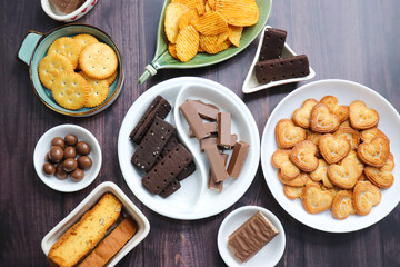 Assorted party snacks on table featuring chocolate cream sandwich biscuits, sugar coated heart cookies, wafer chocolates, chocolate coated caramel balls, salted crackers, and crispy potato chips.
