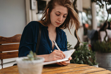 A young woman with light brown hair sits at a wooden table, writing in a notebook with a pen. She is wearing a dark blue shirt and is outdoors in a cafe setting.