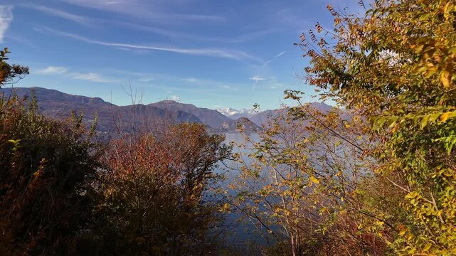 Aerial drone flight over Lake Como with vibrant autumn-winter trees and snow-capped Pre-Alps, capturing colorful foliage and serene alpine reflections in a cinematic lakeside view