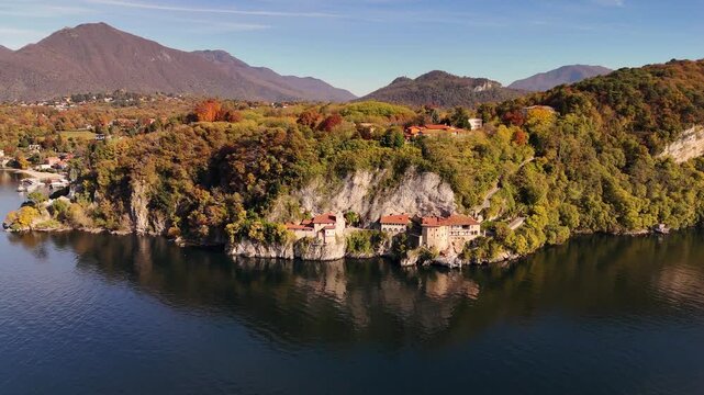 Drone flight over Santa Caterina del Sasso, a cliff-carved monastery with terracotta roofs, surrounded by vibrant autumn colors above Lake Maggiore in Piedmont, Italy