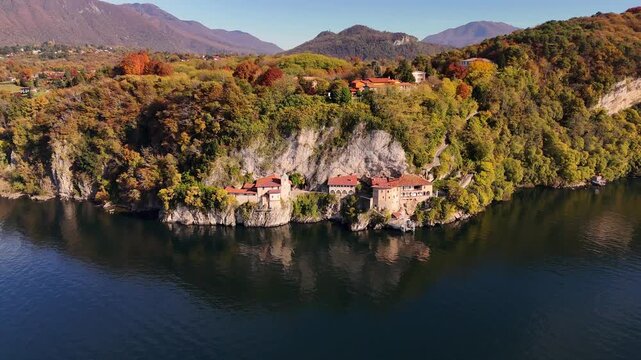 Drone flight over Santa Caterina del Sasso, a cliff-carved monastery with terracotta roofs, surrounded by vibrant autumn colors above Lake Maggiore in Piedmont, Italy