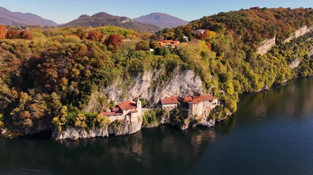 Drone flight over Santa Caterina del Sasso, a cliff-carved monastery with terracotta roofs, surrounded by vibrant autumn colors above Lake Maggiore in Piedmont, Italy