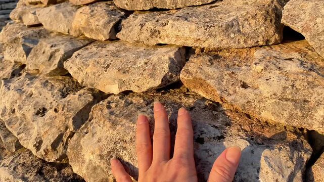  A close-up horizontal shot of a human hand touching the rough, irregular surface of a dry-stone wall or the base of a traditional structure like an Italian trullo. 