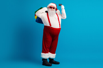 Santa in a bright red costume with suspenders and hat poses with colorful bags and a card against a blue background for a festive Christmas season shopping vibe