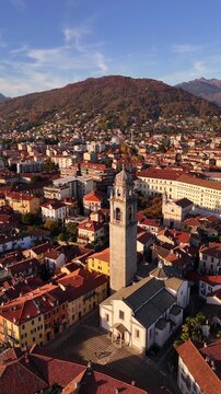 Aerial orbit of a historic stone bell tower rising above an Italian Lake Maggiore village, framed by red rooftops, autumn colors and alpine mountains in a serene cinematic atmosphere