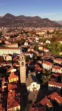 Aerial orbit of a historic stone bell tower in an Italian lakeside village on Lake Maggiore, surrounded by red rooftops and autumn mountains in a soft cinematic evening atmosphere