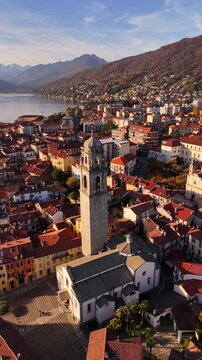 Aerial orbit of a historic stone bell tower in an Italian lakeside village on Lake Maggiore, surrounded by red rooftops and autumn mountains in a soft cinematic evening atmosphere