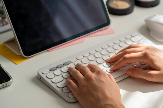 Hands typing on wireless keyboard next to tablet on white desk