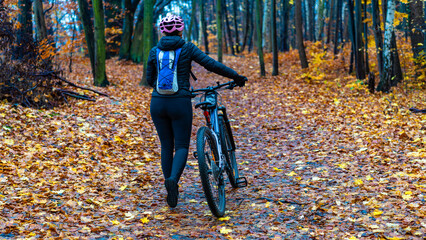 Middle-aged woman riding bicycle on path in forest in autumn. Back view