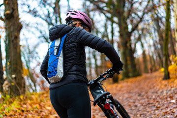 Middle-aged woman riding bicycle on path in forest in autumn. Back view