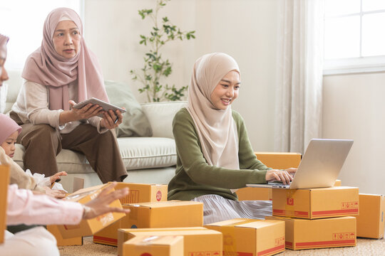 Muslim family running small online business from home, woman using laptop to manage orders surrounded by parcels. Concept of e commerce, home based startup and digital entrepreneurship.