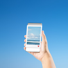 Human hand holding a cellphone displaying a scenic sea view with a blue sky background. Concept of travel, technology, and social media sharing