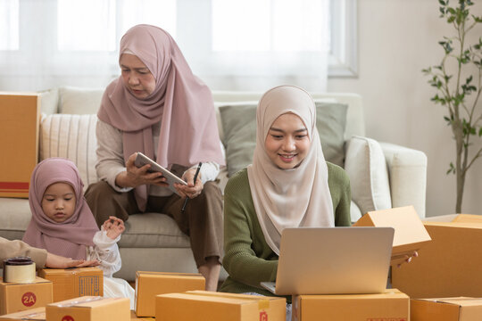 Muslim women working on laptop for online business, surrounded by parcels, with family support. Concept of e-commerce, remote work, female entrepreneurship and small family startup.