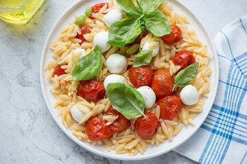 Orzo pasta with mini mozzarella, roasted cherry tomatoes and basil, horizontal shot on a white stone background, elevated view