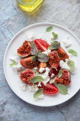 White plate with tomato, cottage cheese and figs salad, vertical shot on a blue and beige stone background, elevated view