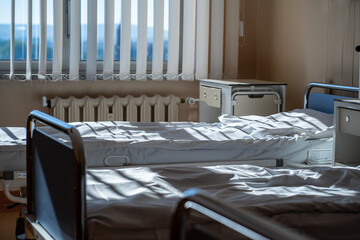 Multiple empty hospital beds with clean sheets lit by morning sunlight in ward